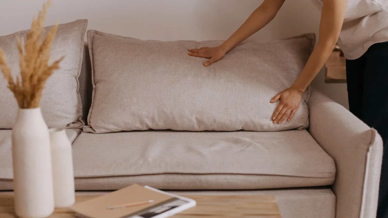 Minimalistic living room scene with a person arranging a pillow on a beige sofa.