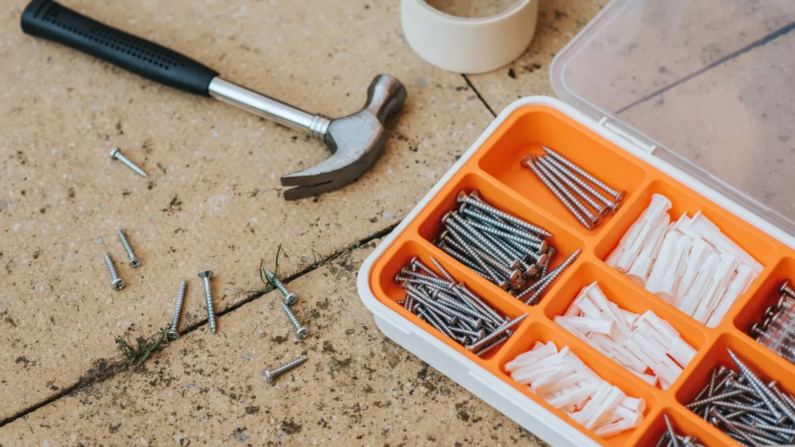 Top view of assorted repair instruments and screws in box near hammer and scotch tape on dirty tile in soft daylight