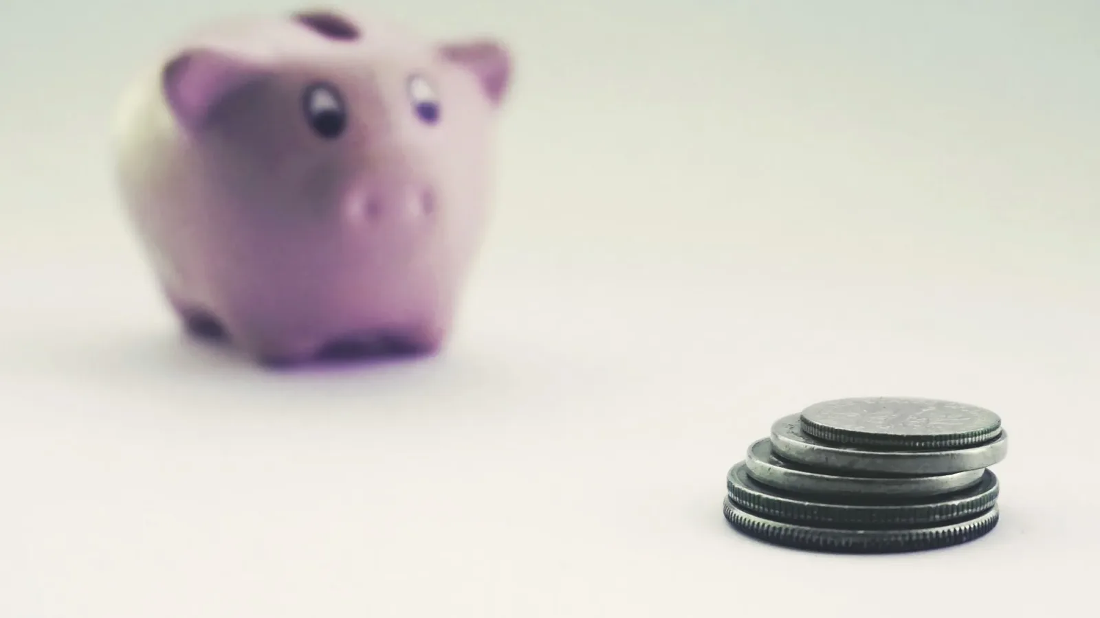 A pink piggy bank blurred in the background with stacked coins in the foreground on a white surface.