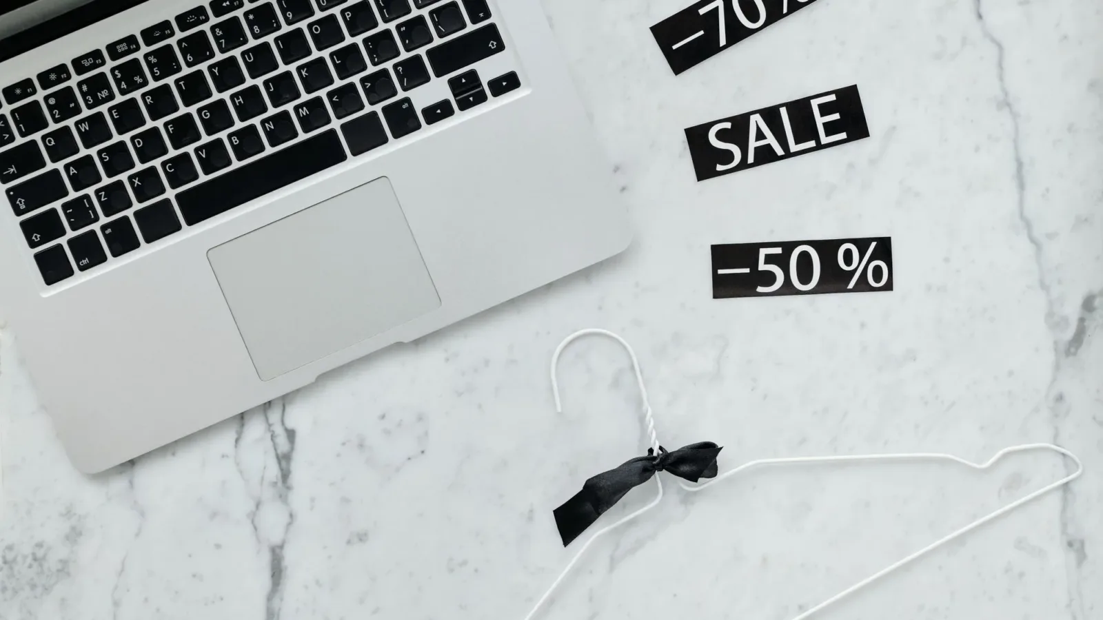 Flat lay of a laptop, hanger, and sale stickers on a marble desk, ideal for Black Friday themes.