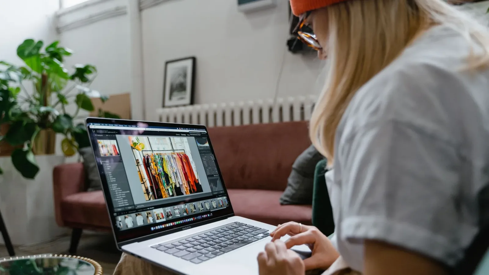 Woman using a laptop for photo editing in a cozy, plant-filled living room setting.